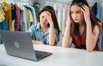 Duas mulheres empreendedoras sentadas em frente a um laptop, com expressões de frustração, analisando os resultados ruins de vendas em sua loja de roupas online.